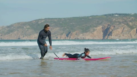 Long shot of happy girl standing on surfboard with help of coach. Slow motion of male instructor with artificial leg wearing diving suit teaching surfing in ocean. Surfing, extreme sport concept