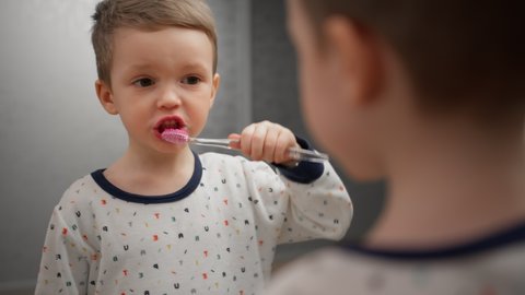 Blonde kid in white pajamas brushing his teeth with toothbrush looks in mirror standing in bathroom. Child is engaged in oral hygiene, fight against caries, brushing teeth from plaque and pollution.