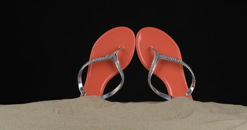Pair of pink flip flops embellished with rhinestones stick out of the sand. Panorama. Isolated on black background