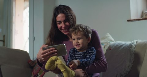 Cinematic shot of happy smiling mother and her cute toddler baby boy having fun to use technology smart phone together for family entertainment on sofa in living room at home.