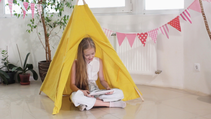 A cute little girl sitting cross-legged on the floor in a yellow wigwam watching cartoons on a tablet thoughtfully lifting her head up