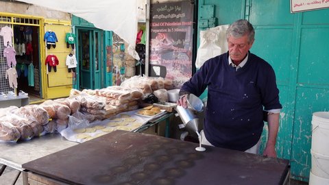BETHLEHEM, WEST BANK – NOVEMBER 5 2021: Senior Palestinian man pours dough on hot plate, making fresh pancakes in outdoor bakery on market in Bethlehem, West Bank
