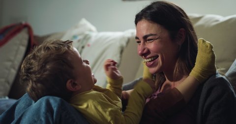 Cinematic authentic close up shot of young happy smiling mother is tickling and playing with her toddler baby boy son on sofa in living room at home.
