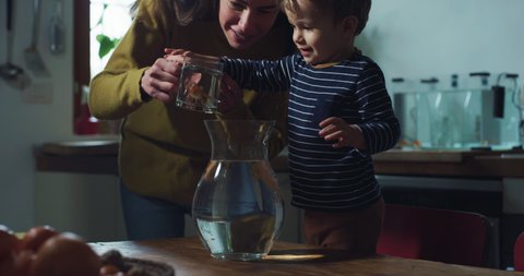 Cinematic shot of young mother puts domestic goldfishes in fish bowl and explains to her curious happy little toddler boy how to feed and take care of them in kitchen at home.