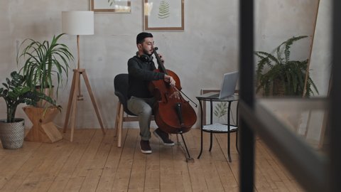 Wide tracking left of young Asian male professional instrumentalist wearing headphones around neck, playing double bass on computer camera indoors at daytime