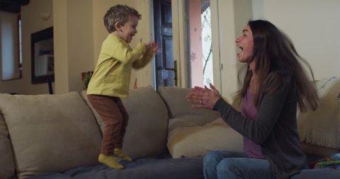 Cinematic authentic shot of young happy smiling mother is having fun to dance together with her toddler baby boy son while watching television on sofa in living room at home.