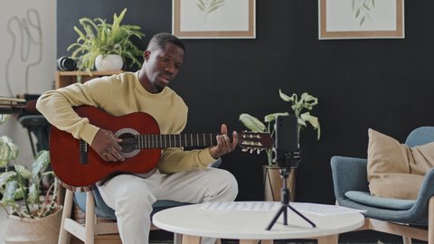 Medium long of young Black man sitting in armchair at home at daytime, playing acoustic guitar, smiling and talking on smartphone camera, streaming video online