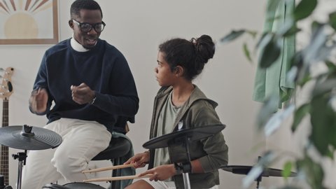 Medium long of preppy Black male tutor wearing eyeglasses, teaching eleven-year-old African American girl use drumsticks on e-drum in modern apartment at daytime