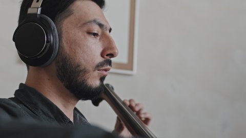 Low angle side view of young Asian male professional musician wearing earphones playing double bass indoors at daytime