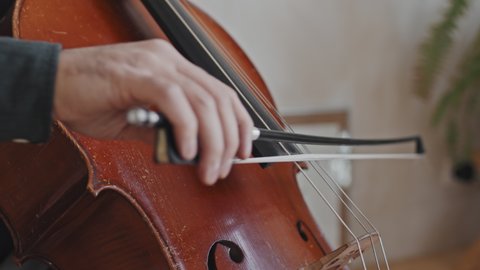 Low angle side view of cropped male musician slowly playing double bass with bow indoors at daytime