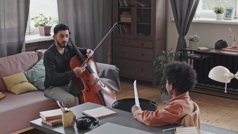 Medium long of formally dressed Asian man sitting on couch in apartment at daytime, playing double bass, little Black boy watching and learning
