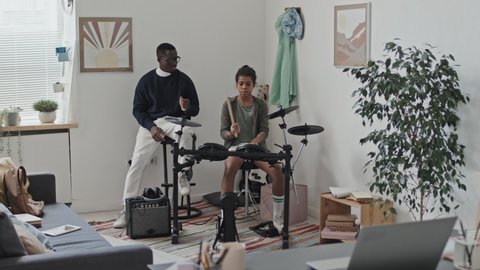 Wide shot of young Black male teacher watching eleven-year-old girl playing electronic drum kit in modern studio at daytime, smiling