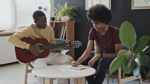 Medium long of curly-haired young man sitting in armchair in living room at daytime, taking notes when Black male music teacher showing chords on acoustic guitar and talking