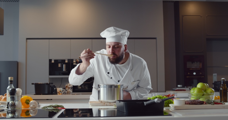 Professional chef tasting sauce and showing sign for delicious. Portrait of handsome male cook in uniform preparing soup in restaurant kitchen and doing taste approval gesture