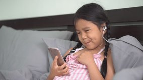 Asian child or kid girl wearing headphones or small talk to study online or learn from home to listening music on smartphone or play mobile phone for learning to sitting relax on bed and lean pillow
