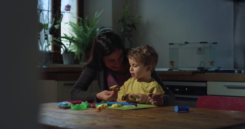 Cinematic shot of young mother teaching and playing to her curious happy little toddler boy how to sculpt from clay and play while playing together in kitchen at home.