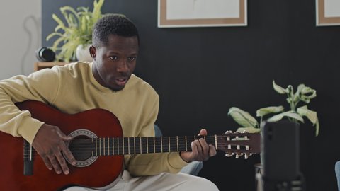 Medium of young Black man sitting in armchair at home at daytime, playing acoustic guitar, smiling and talking on smartphone camera, streaming live video