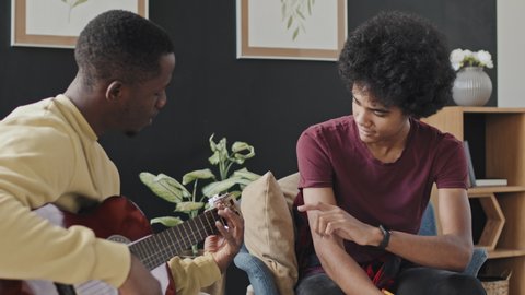 Tilting up of curly-haired young man sitting in armchair at home at daytime, taking notes when Black male music tutor playing acoustic guitar and talking