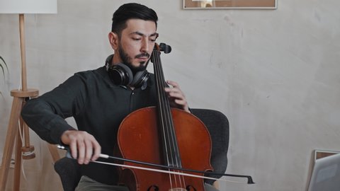Tilting down of young Asian male professional instrumentalist wearing headphones around neck, playing double bass on computer camera indoors at daytime