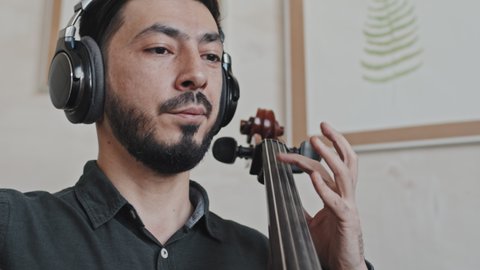 Low angle of young Asian male professional musician wearing headphones playing double bass indoors at daytime