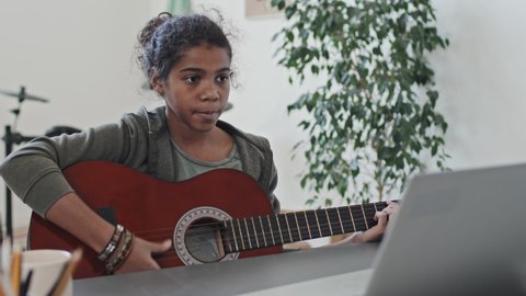 Medium close-up of eleven-year-old Black girl having online acoustic guitar lesson via portable computer in her room in modern apartment at daytime