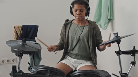 Tilting down of talented eleven-year-old African American girl wearing over-ear headphones, enjoying playing e-drum in her room modern apartment at daytime