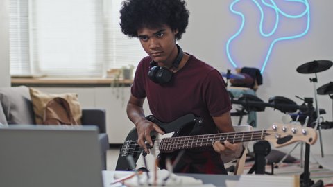 Medium of young curly-haired Black man wearing headphones around neck, sitting at desk in his room at daytime, learning to play bass guitar, watching tutorial on laptop computer