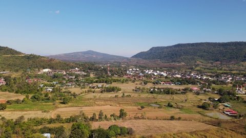 Aerial footage sliding towards the left revealing a town in a valley with beautiful mountains and blue sky near Wat Somdet Phu Ruea, Ming Mueang, Loei in Thailand.