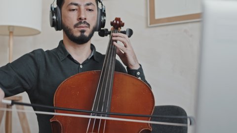 Tilting up and down of young Asian man wearing over-ear headphones and beard, sitting in armchair at home, playing double bass on portable computer camera