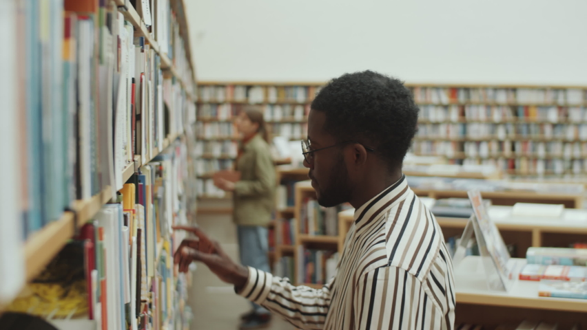 African American man in glasses and striped shirt taking book from shelf and reading it in library