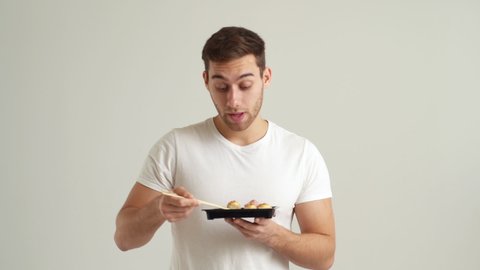 Studio portrait of handsome young man eating fresh delicious sushi rolls with chopsticks on white isolated background. Happy Caucasian male eats Asian food with closed eyes. Shooting in slow motion.