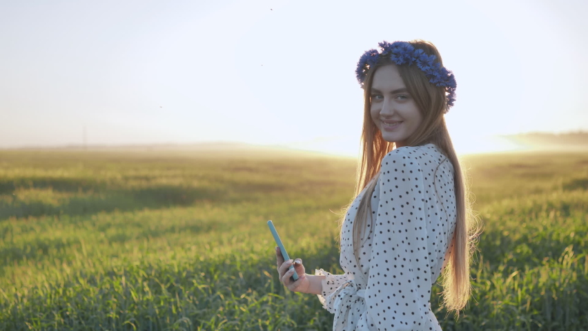 A cute Ukranian girl takes selfies in a field with a wreath on her head.