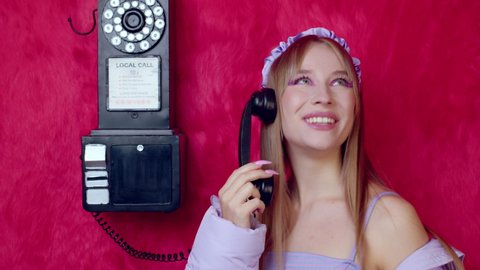 Coquettish vintage girl with bright makeup talking on retro wall phone. Young blonde lady chatting with boyfriend or soulmate in room with pink fur wallpaper. Sharing latest news, gossip