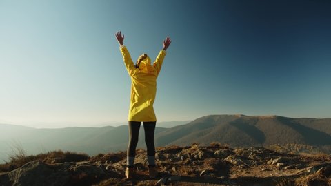 Camera Follows Young Woman In Yellow Jacket Running Up On Top Of Mountain Raises Arms Into Air, Happy And Drunk On Life And Happiness. Cheerful Girl Exploring Nature Happy. Admires Rising Sun, Sunset