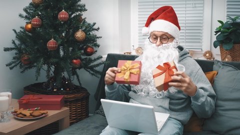 A man in a Santa Claus costume with a beard shows gifts via video link on a laptop for Christmas to his relatives and friends, sitting at home on the couch next to the Christmas tree.