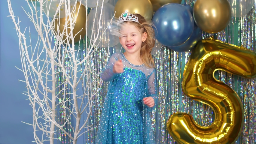 Happy child dancing and having fun. white and blue balloons fall from above. A girl waves a magic wand and makes a wish in a dress and with a crown. Little princess on birthday. 5 years and tinsel.