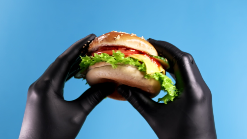 Hands in black latex gloves with burger on blue background. Man demonstrating appetizing patty with meat cutlet, vegetables, melted cheese and mayonnaise. Fast food concept.