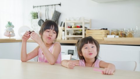 Asian two sweet sisters little girl enjoy eat fast food, potato chips. Hungry preschool girl kid sit on table in kitchen puts snack in mouth with hand. Tasty and unhealthy food for children concept.