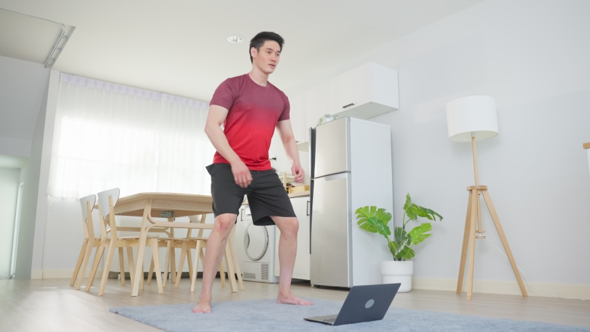 Asian young handsome man doing cardio exercise in living room at home. Attractive active male doing squatting workout on floor follow instruction video from online trainer on laptop computer in house. Royalty-Free Stock Footage #1086268499