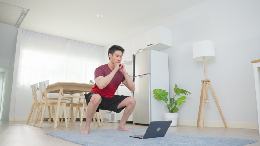 Asian young handsome man doing cardio exercise in living room at home. Attractive active male doing squatting workout on floor follow instruction video from online trainer on laptop computer in house. Royalty-Free Stock Footage #1086268499