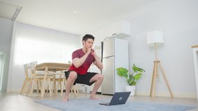 Asian young handsome man doing cardio exercise in living room at home. Attractive active male doing squatting workout on floor follow instruction video from online trainer on laptop computer in house.