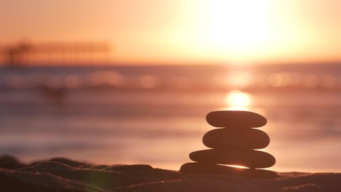 Stack of pebble stones, sandy ocean beach, sunset sky. Rock balancing in sun light, sea water waves. Stones staking in pyramid pile. Zen meditation and harmony in balance. Seamless looped cinemagraph.
