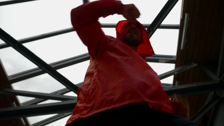 a man in a red jacket with a hood is boxing against the background of iron structures. bottom view