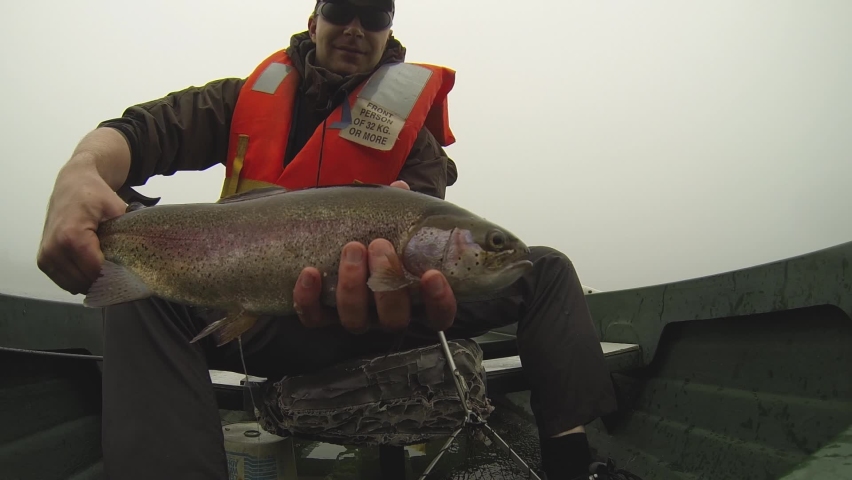 A fly fishing man holding a rainbow trout which he has just caught, while sitting in a boat
