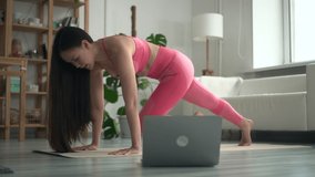 Young woman engaged in fitness and talking in front of laptop in home room spbd. Close-up view of beautiful Asian female does exercise on floor and looks at computer screen, tells and shows how to do
