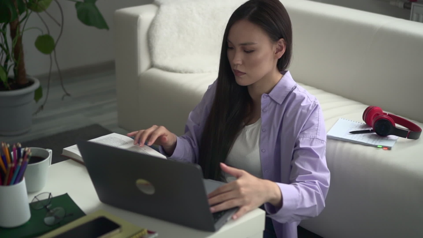 Young businesswoman or student typing in front of laptop and sitting at table in apartment spbd. Beautiful Asian woman looks at computer screen and types text, reads book or notebook and sits at desk
