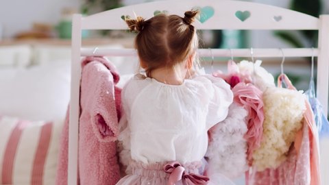 rear view of cute little girl, fashionista decides what to wear standing in front of the clothing rack stand at sunny room