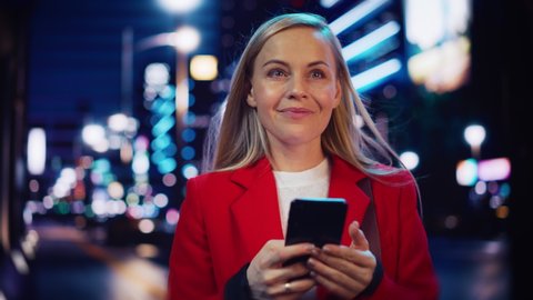 Beautiful Woman Standing, Using Smartphone on City Street with Neon Bokeh Lights Shining at Night. Confident Smiling Beautiful Female Using Mobile Phone. Medium Tracking Cinematic Portrait.