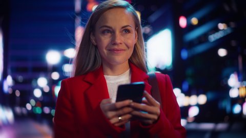 Beautiful Woman Standing, Using Smartphone on City Street with Neon Bokeh Lights Shining at Night. Confident Smiling Beautiful Female Using Mobile Phone. Medium Tracking Cinematic Portrait.