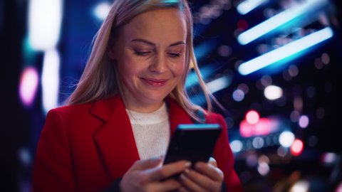 Close Up Portrait of a Beautiful Woman in Red Coat Walking in a Modern City Street with Neon Lights at Night. Attractive Female Using Smartphone and Looking Around the Urban Cinematic Environment.